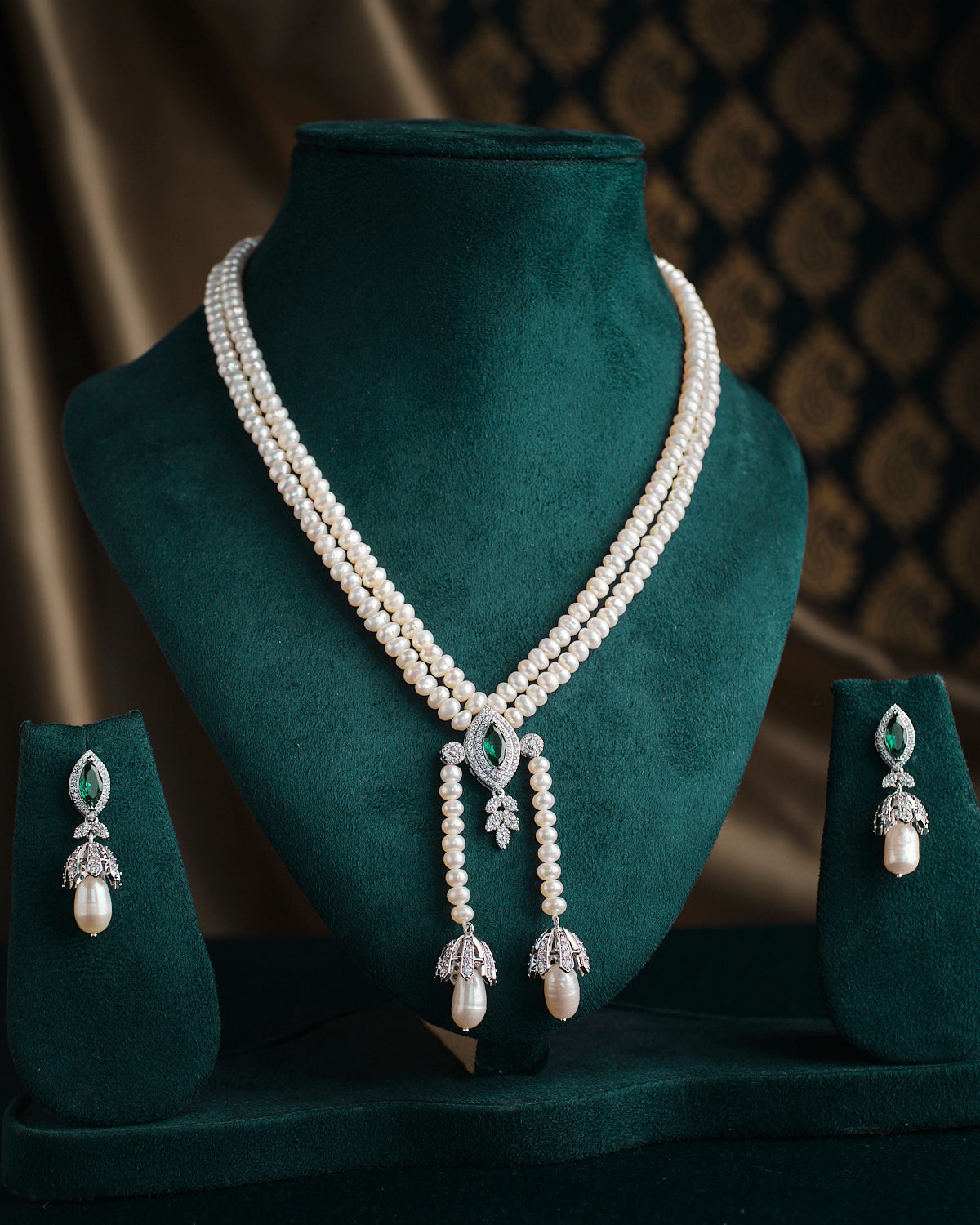 The Kanaka Mridula Pearl Necklace Set by Chandrani Pearls, featuring a necklace with pearls and green gemstones along with matching earrings, elegantly presented on a green velvet stand.