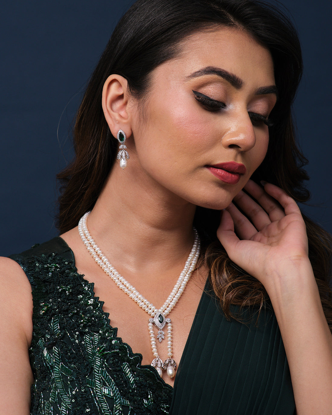 The Kanaka Mridula Pearl Necklace Set by Chandrani Pearls, featuring a necklace with pearls and green gemstones along with matching earrings, elegantly presented on a green velvet stand.