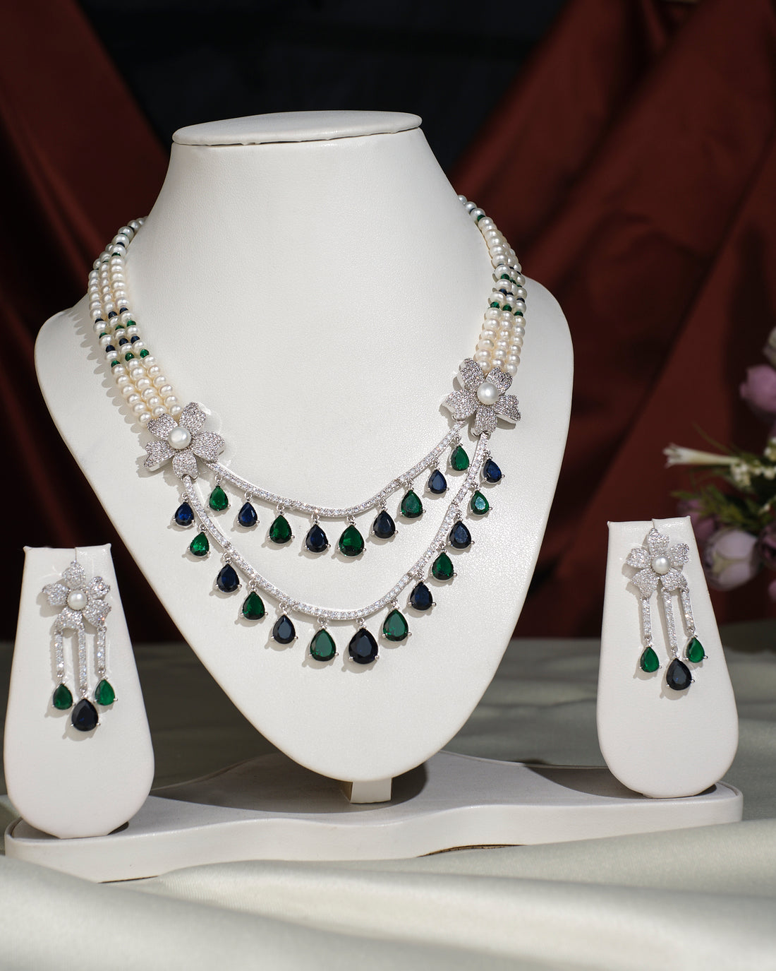 The Beautiful Ravishing Pearl Necklace Set by Chandrani Pearls India, featuring pearls, diamonds, and emeralds with matching earrings, is showcased on a white jewelry stand against a neutral background.