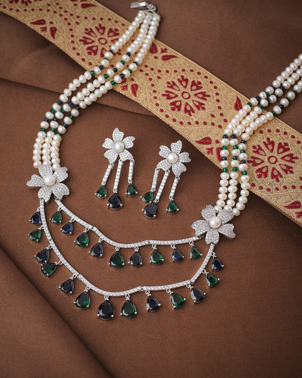 The Royal Legacy Pearl CZ Necklace Set by Chandrani Pearls features pearl and CZ brilliance with green and blue gemstone drops and matching floral earrings, displayed elegantly on a brown and red patterned fabric.