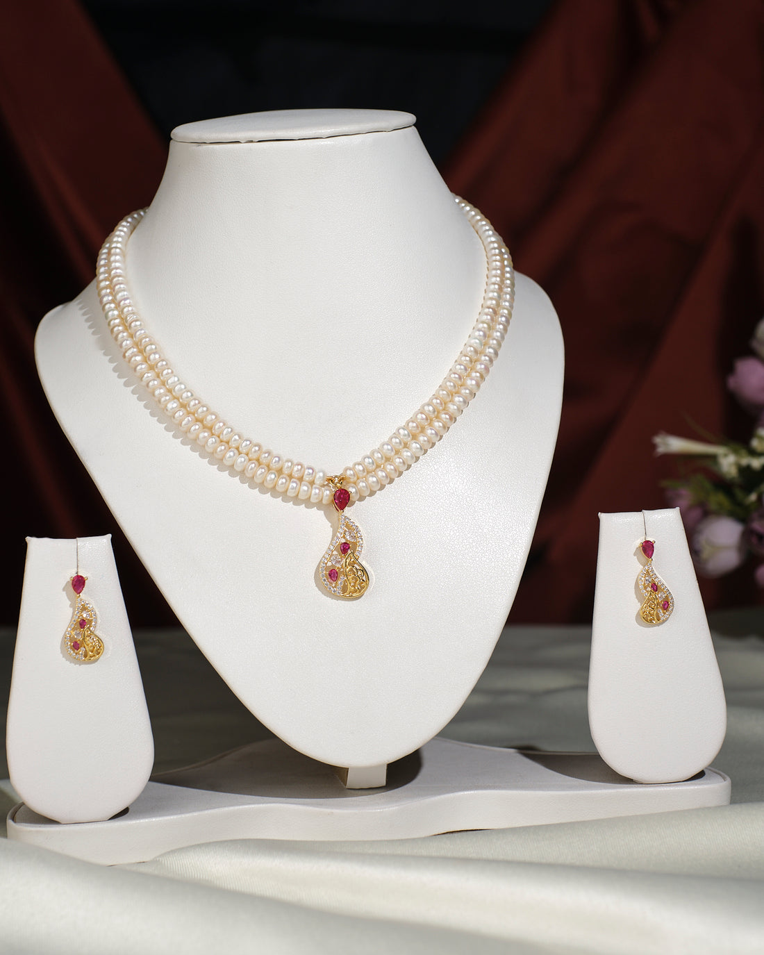 The Elegant Pearl Necklace Set by Chandrani Pearls India features lustrous pearls and a gold-red pendant, elegantly displayed with matching earrings on a stand against a dark backdrop.