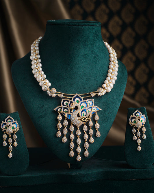 The Mayura Dripping Elegance Pearl Necklace Set by Chandrani Pearls features a sophisticated combination of pearls and gold, adorned with an intricate blue and gold peacock design. The set includes matching earrings and is elegantly displayed on a teal velvet stand.