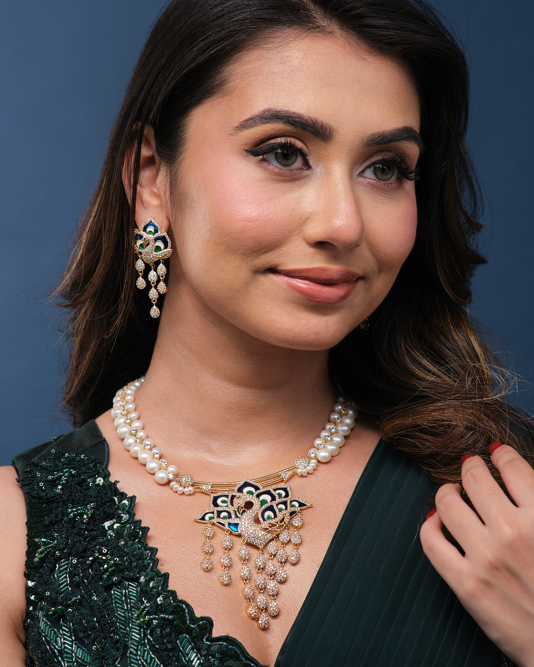 The Mayura Dripping Elegance Pearl Necklace Set by Chandrani Pearls features a sophisticated combination of pearls and gold, adorned with an intricate blue and gold peacock design. The set includes matching earrings and is elegantly displayed on a teal velvet stand.