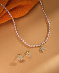 The Rosé Royale Necklace Set by Chandrani Pearls features a pearl necklace with a gold and diamond floral pendant and matching stud earrings, beautifully displayed on a tan surface with orange fabric—ideal for wedding jewelry.