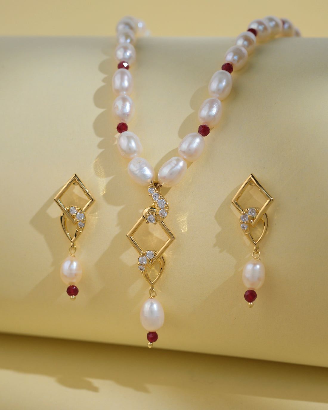 The Regal 1 Line Necklace Set by Chandrani Pearls India features a delicate necklace and matching earrings with gold geometric shapes, pearls, red beads, and clear stones, beautifully displayed on a pale yellow surface.