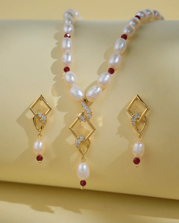 The Regal 1 Line Necklace Set by Chandrani Pearls India features a delicate necklace and matching earrings with gold geometric shapes, pearls, red beads, and clear stones, beautifully displayed on a pale yellow surface.