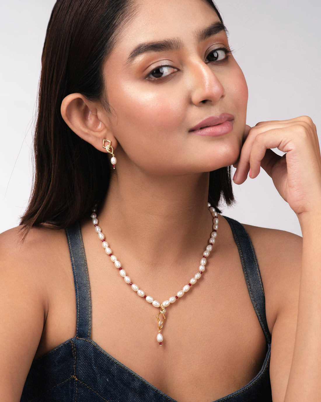 The Regal 1 Line Necklace Set by Chandrani Pearls India features a delicate necklace and matching earrings with gold geometric shapes, pearls, red beads, and clear stones, beautifully displayed on a pale yellow surface.