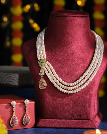 The Vividhruma - Colourful Blossoms Necklace Set by Chandrani Pearls, featuring a white pearl necklace with gold and gem accents, is showcased on a red stand. Matching earrings rest on a red box against a backdrop of blurred yellow and red decorations.