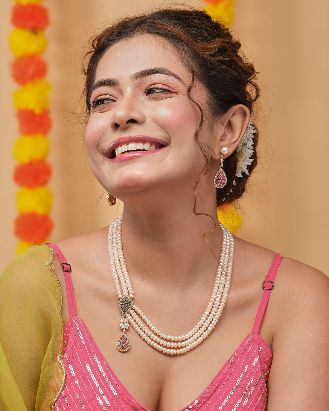 The Vividhruma - Colourful Blossoms Necklace Set by Chandrani Pearls, featuring a white pearl necklace with gold and gem accents, is showcased on a red stand. Matching earrings rest on a red box against a backdrop of blurred yellow and red decorations.