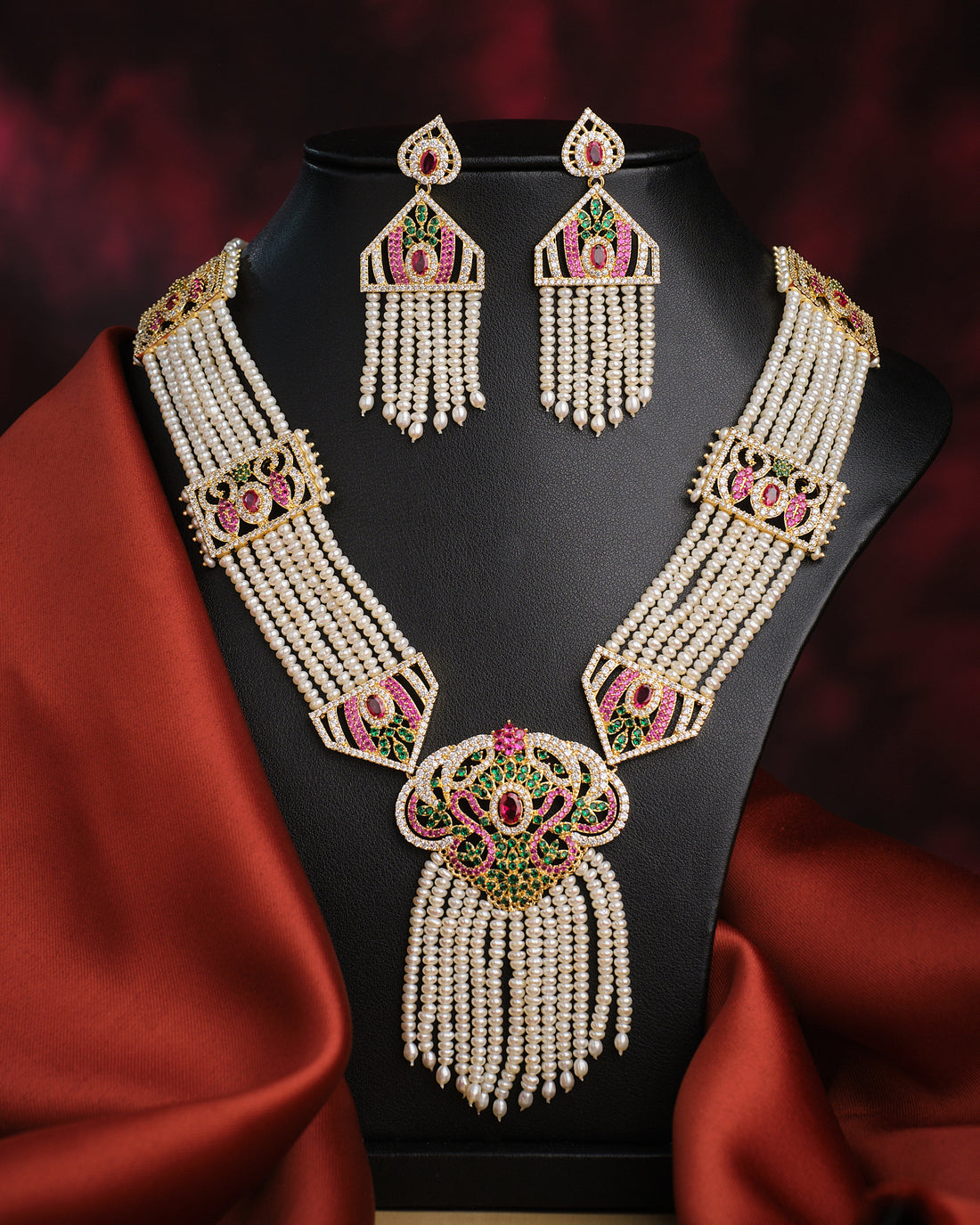 The Sindurya-Scarlet Ray's Lahari Sitahar Set by Chandrani Pearls features a multi-strand pearl necklace with matching earrings, gold, green, and pink accents, vintage-style clasp—displayed on black against red fabric—a timeless heirloom piece.