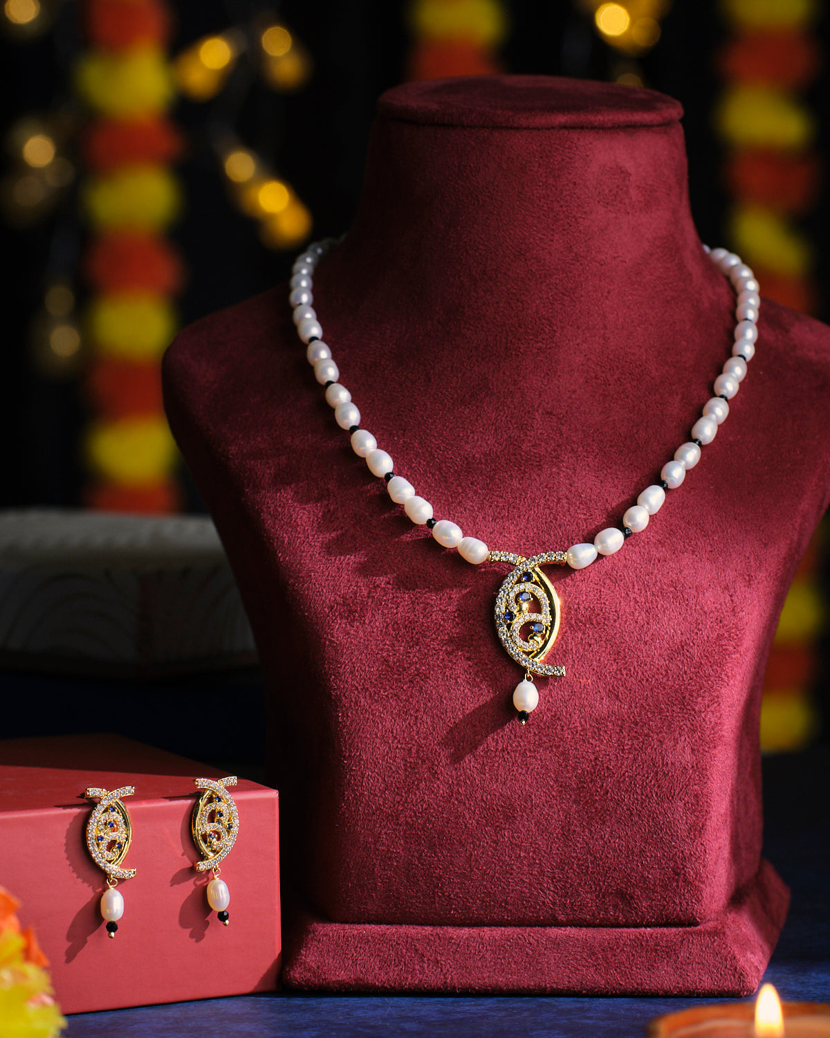 The Adorable Aura Pearl Necklace Set by Chandrani Pearls, featuring a necklace with a decorative pendant elegantly displayed on a velvet stand, is complemented by matching earrings presented on a red box, all set against a softly lit background.