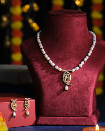 The Adorable Aura Pearl Necklace Set by Chandrani Pearls, featuring a necklace with a decorative pendant elegantly displayed on a velvet stand, is complemented by matching earrings presented on a red box, all set against a softly lit background.