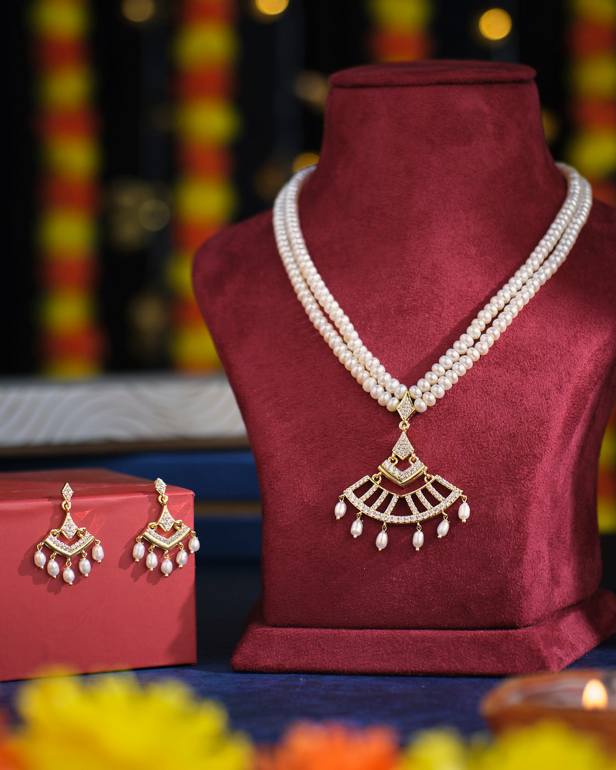 The Eternal Treasure Pearl Necklace Set by Chandrani Pearls, featuring a gold pendant, is displayed on a maroon stand. Matching earrings accompany the set, elegantly presented on a red box.