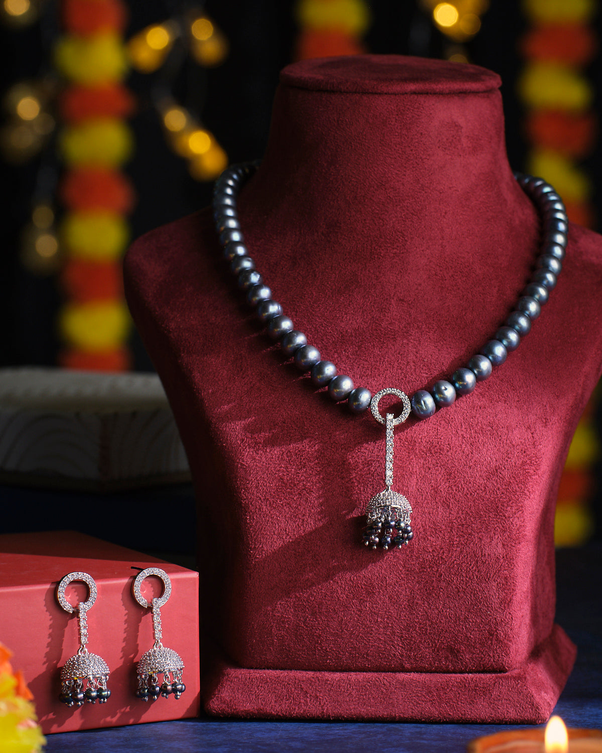 The Mystic Midnight Pearl Necklace Set by Chandrani Pearls is displayed on a red velvet stand, featuring a dangling pendant and accompanied by matching earrings placed on a nearby red box.