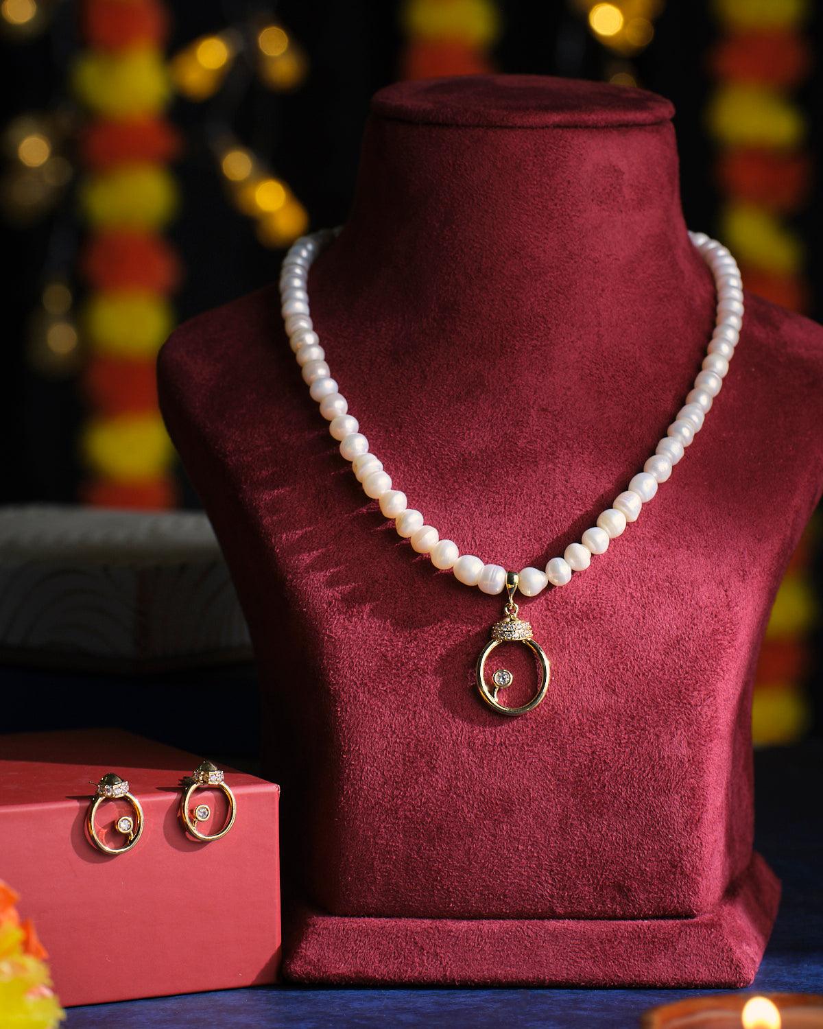 The Queen Crown Pearl Necklace Set by Chandrani Pearls, featuring a luxurious pearl necklace with a gold pendant, elegantly displayed on a burgundy stand and accompanied by a red box containing exquisite gold earrings.