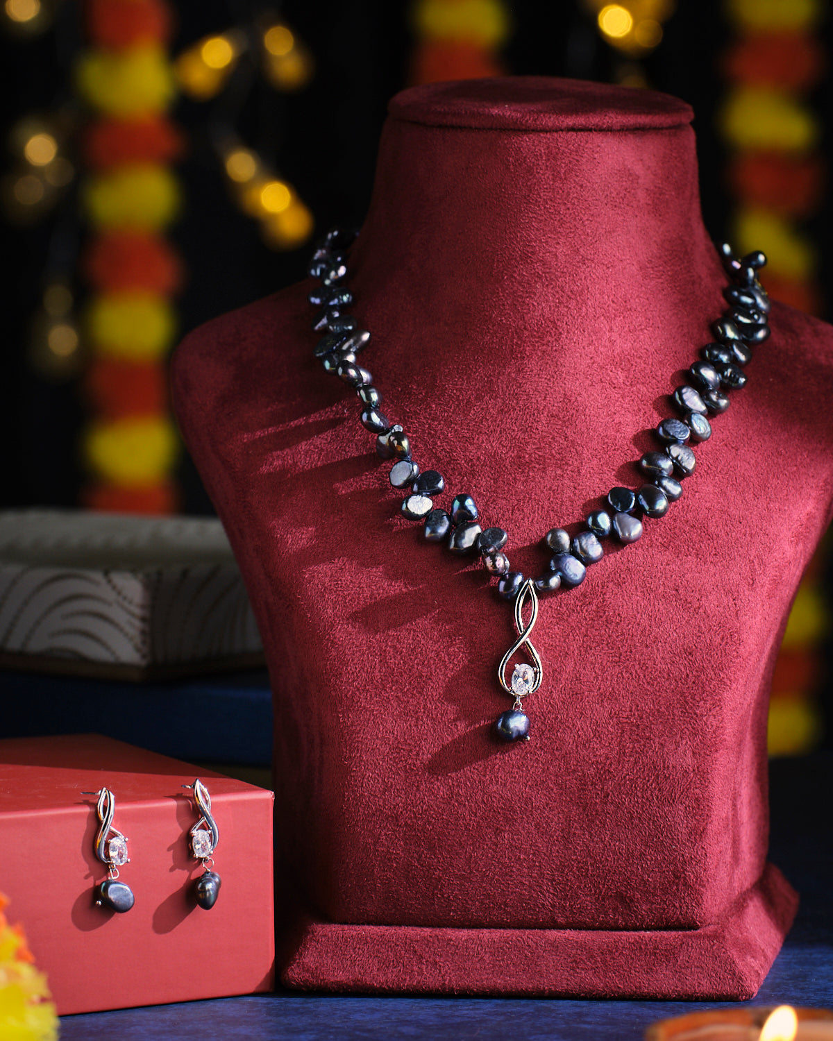 The Vashikrit - The Hypnotic Tiers Pearl Necklace Set by Chandrani Pearls is elegantly displayed on a red velvet bust, with matching earrings showcased on a small red box.