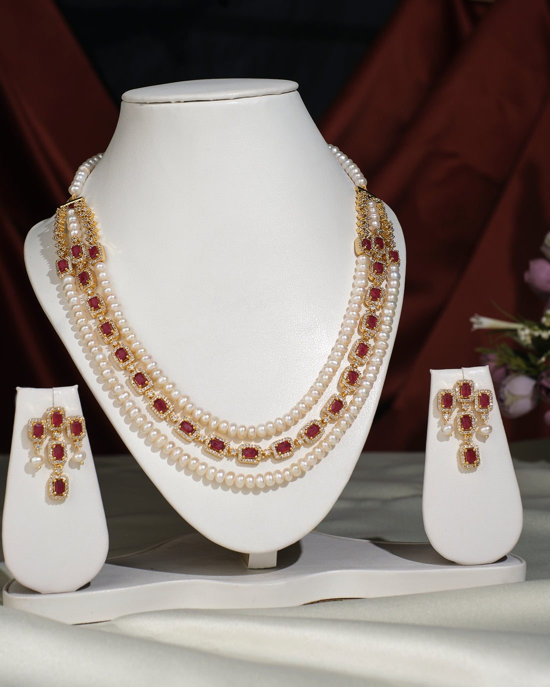 The Gorgeous Pearl Necklace Set by Chandrani Pearls India, featuring red and gold accents and matching earrings, is displayed on a white stand against draped fabric—perfect for both formal and casual looks.