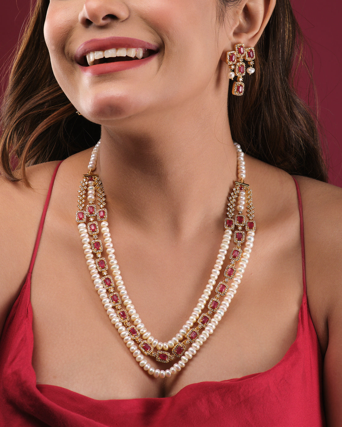 The Gorgeous Pearl Necklace Set by Chandrani Pearls India, featuring red and gold accents and matching earrings, is displayed on a white stand against draped fabric—perfect for both formal and casual looks.