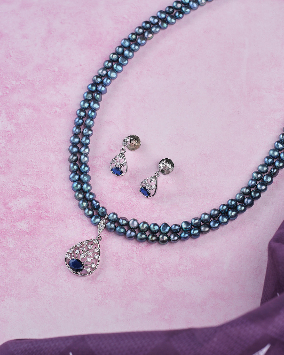 The Trendy Real Pearl Necklace Set by Chandrani Pearls features double strands with a stone-studded pendant and matching teardrop earrings, perfect for parties, shown on a pink surface.