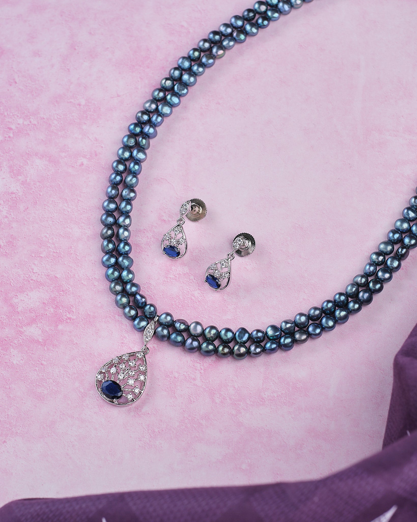 The Trendy Real Pearl Necklace Set by Chandrani Pearls features double strands with a stone-studded pendant and matching teardrop earrings, perfect for parties, shown on a pink surface.