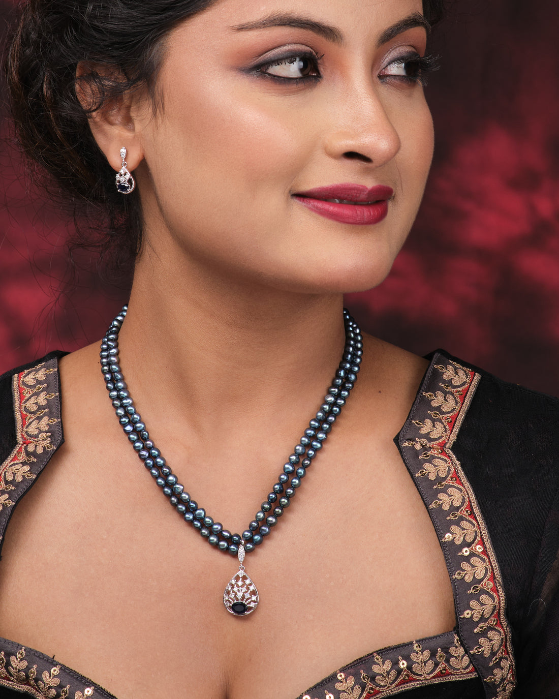 The Trendy Real Pearl Necklace Set by Chandrani Pearls features double strands with a stone-studded pendant and matching teardrop earrings, perfect for parties, shown on a pink surface.