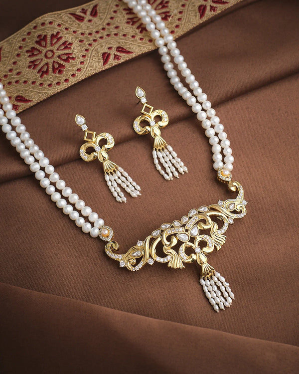 The Imperial Elegance Pearl Necklace Set by Chandrani Pearls, featuring gold, pearls, and shimmering CZ details with matching earrings, is showcased on brown fabric beside a decorative embroidered textile.