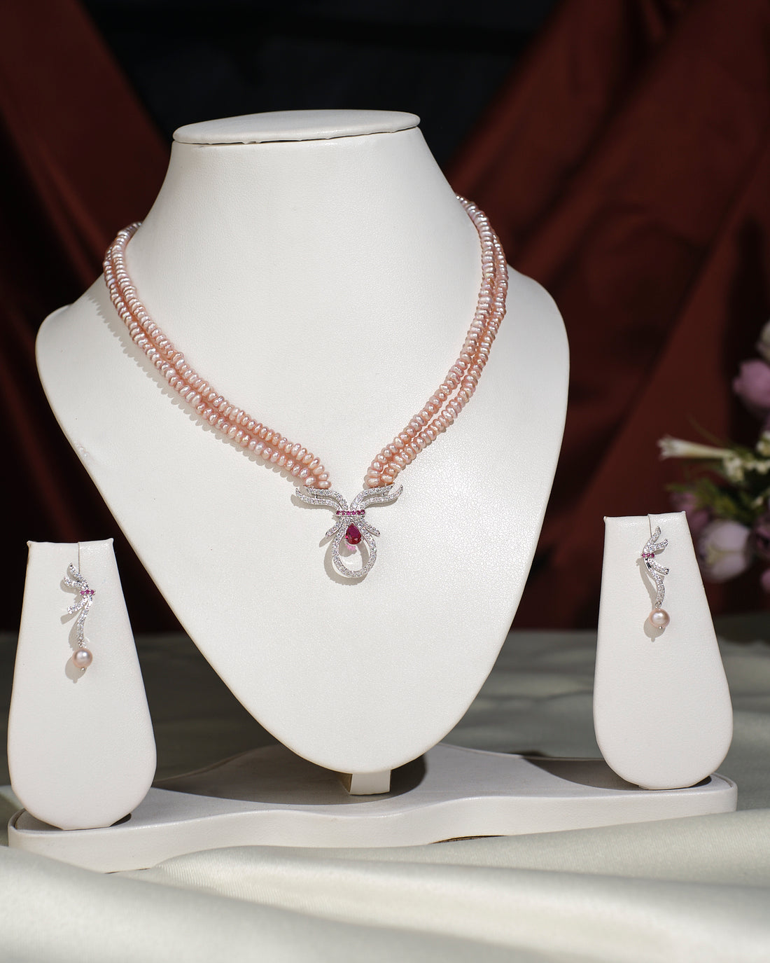 The Beautiful Pearl Necklace Set by Chandrani Pearls India features a pink beaded necklace with pearls and a central pendant, plus matching earrings—ideal for both casual and formal wear. Displayed elegantly on stands against a neutral background.