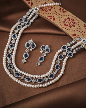 The Noor Royal Light CZ Pearl Necklace Set by Chandrani Pearls includes a pearl and blue gemstone necklace, matching earrings, and a ring with radiant pearls and CZ accents, displayed on brown fabric with a patterned strip.