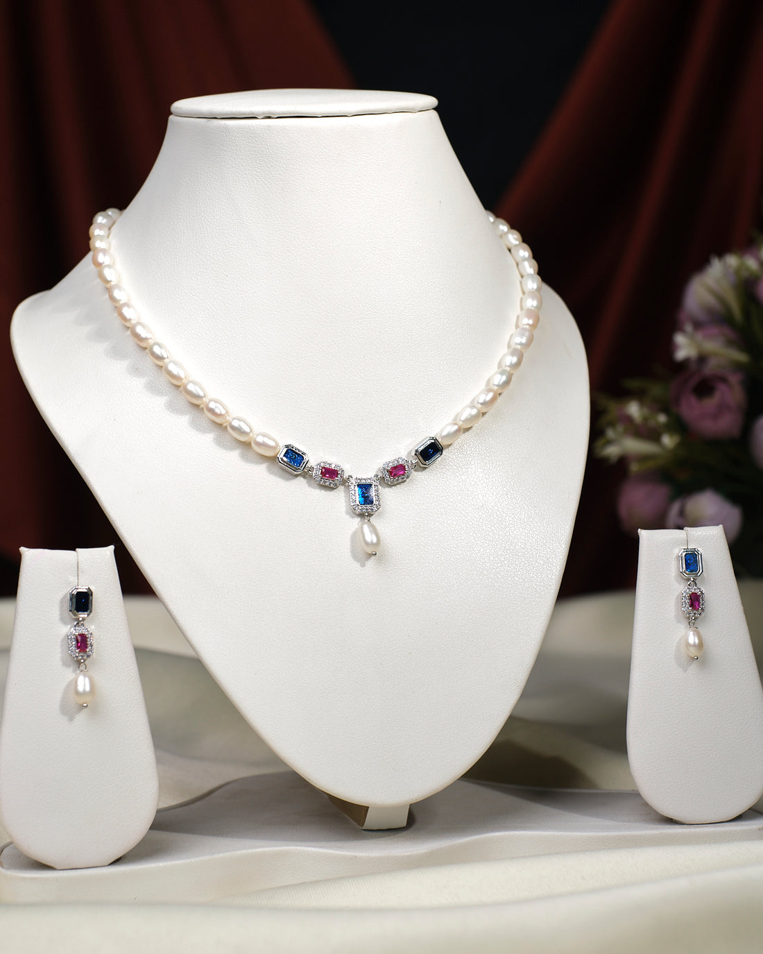 The Camelia CZ Stone 1 Line Necklace Set by Chandrani Pearls India is displayed on a stand, featuring a pearl necklace with blue and pink stones, drop pendant, and matching earrings—an elegant addition to any wardrobe.