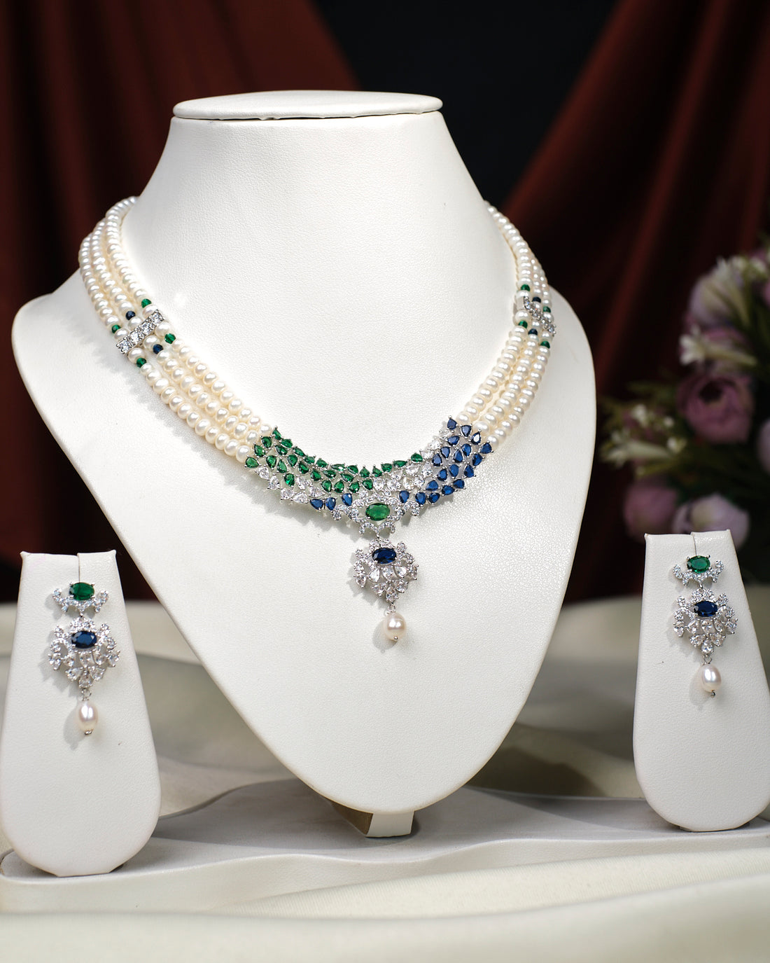 The Crystal Cascade CZ Stone 3 Line Necklace Set by Chandrani Pearls India, featuring green and blue gemstone accents with matching earrings, is displayed on a white stand against a floral backdrop—an elegant addition to any wardrobe.