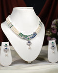 The Crystal Cascade CZ Stone 3 Line Necklace Set by Chandrani Pearls India, featuring green and blue gemstone accents with matching earrings, is displayed on a white stand against a floral backdrop—an elegant addition to any wardrobe.