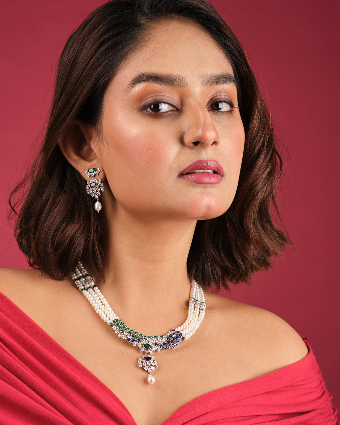 The Crystal Cascade CZ Stone 3 Line Necklace Set by Chandrani Pearls India, featuring green and blue gemstone accents with matching earrings, is displayed on a white stand against a floral backdrop—an elegant addition to any wardrobe.