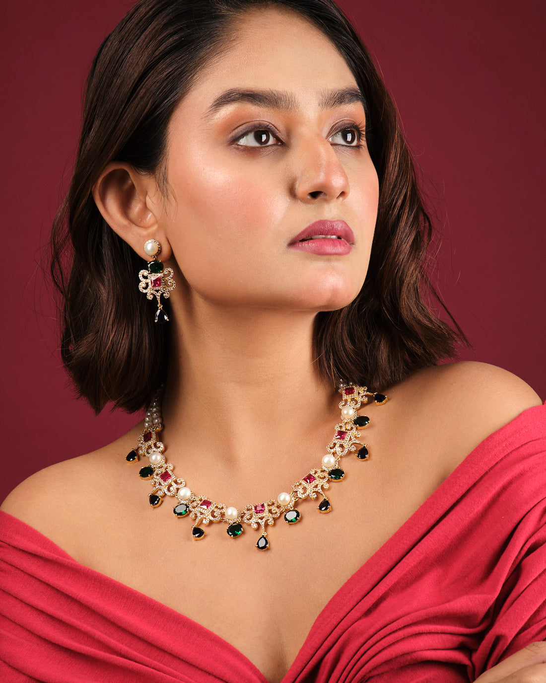 The Mosaic Marvel CZ Stone 2 Line Necklace Set by Chandrani Pearls India features chic gold tones with red, green, and pearl accents, displayed on a white stand alongside matching dangle earrings—an elegant addition to your wardrobe.