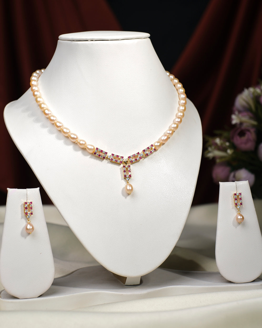 The Yamie CZ Stone 1 Line Necklace Set by Chandrani Pearls India features a pearl pendant necklace and matching drop earrings on display stands, bringing timeless elegance to any wardrobe with a floral backdrop.