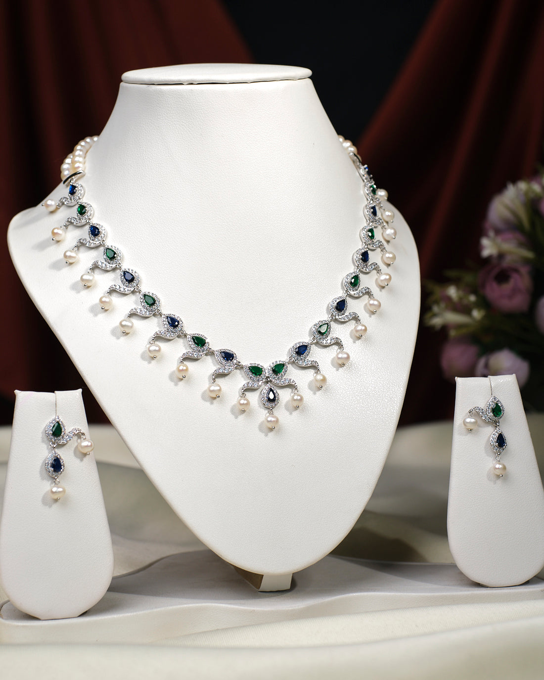 The Asmara CZ Stone Necklace Set by Chandrani Pearls India, featuring a pearl and gemstone necklace with matching earrings, is elegantly displayed on white stands against a floral background.