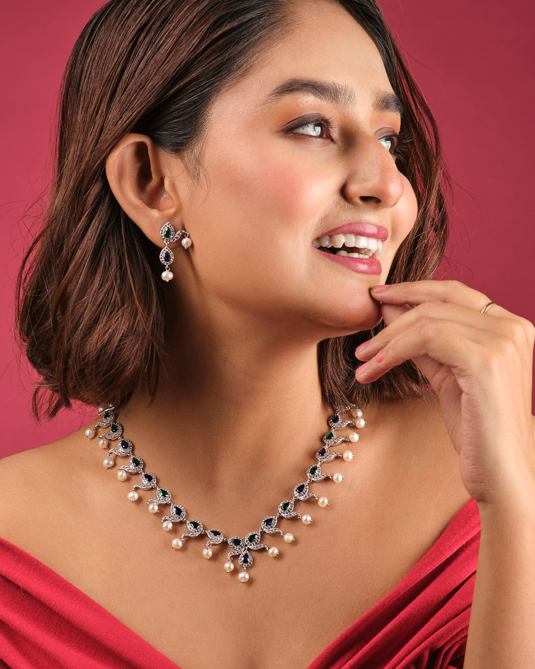 The Asmara CZ Stone Necklace Set by Chandrani Pearls India, featuring a pearl and gemstone necklace with matching earrings, is elegantly displayed on white stands against a floral background.
