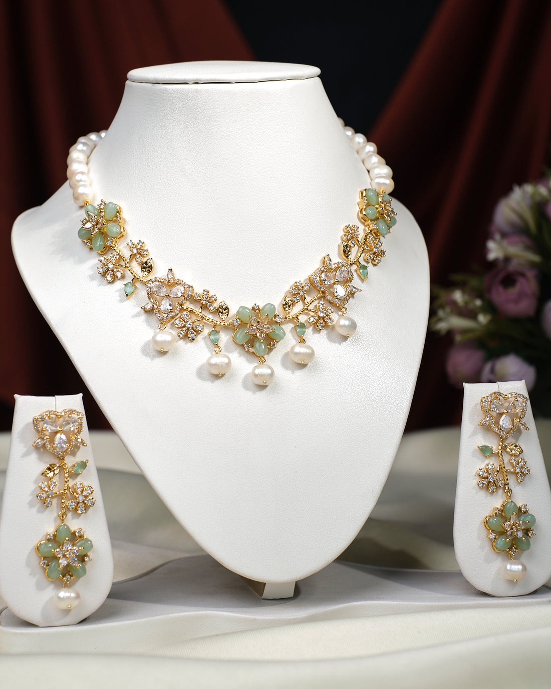 The Sangini Floral Necklace Set by Chandrani Pearls India features gold and pearls with green floral accents, paired with matching earrings, elegantly displayed on a white stand against a dark burgundy backdrop.