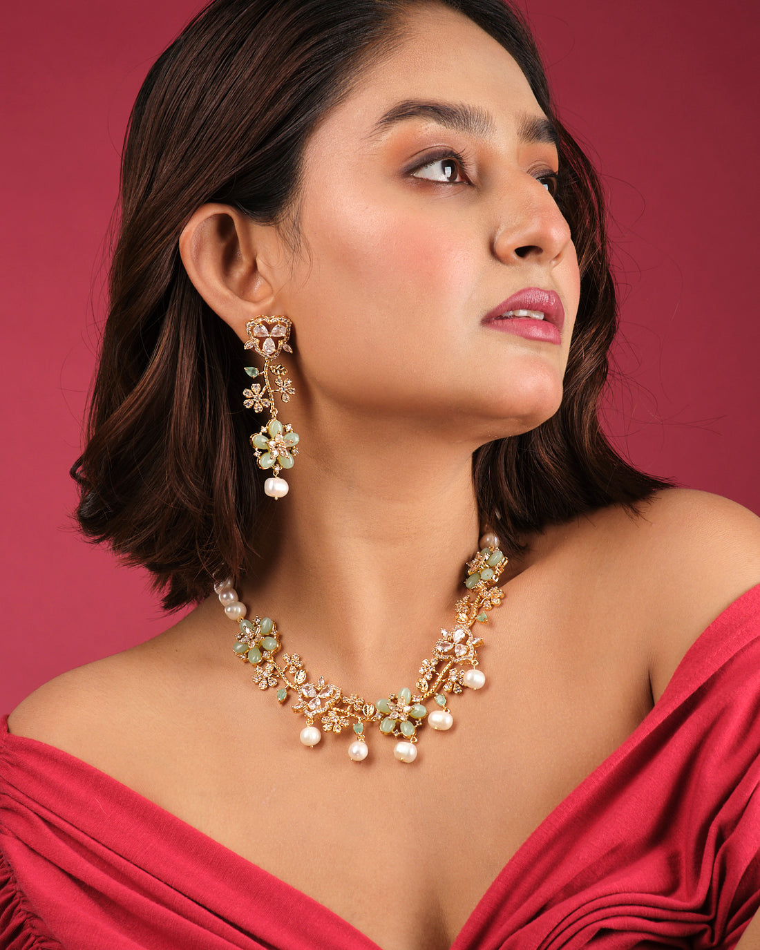 The Sangini Floral Necklace Set by Chandrani Pearls India features gold and pearls with green floral accents, paired with matching earrings, elegantly displayed on a white stand against a dark burgundy backdrop.