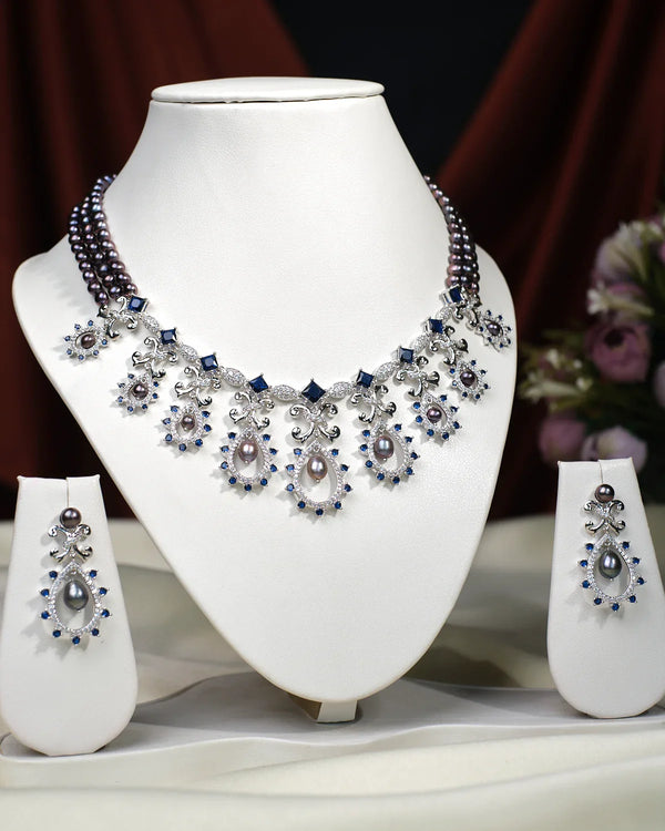 The Gourgeous Sharanya Necklace Set by Chandrani Pearls India, featuring pearls and gemstones with matching earrings, is displayed on a white stand against a blurred floral background.