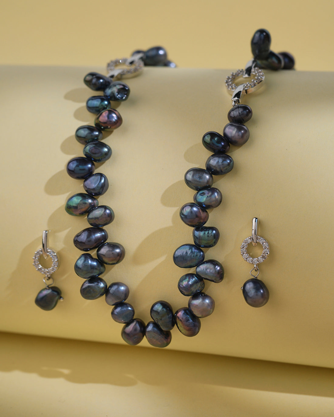 The Trendy Pearl Necklace Set by Chandrani Pearls India features dark pearls and small circular diamond accents, exuding timeless brilliance against a light yellow background.
