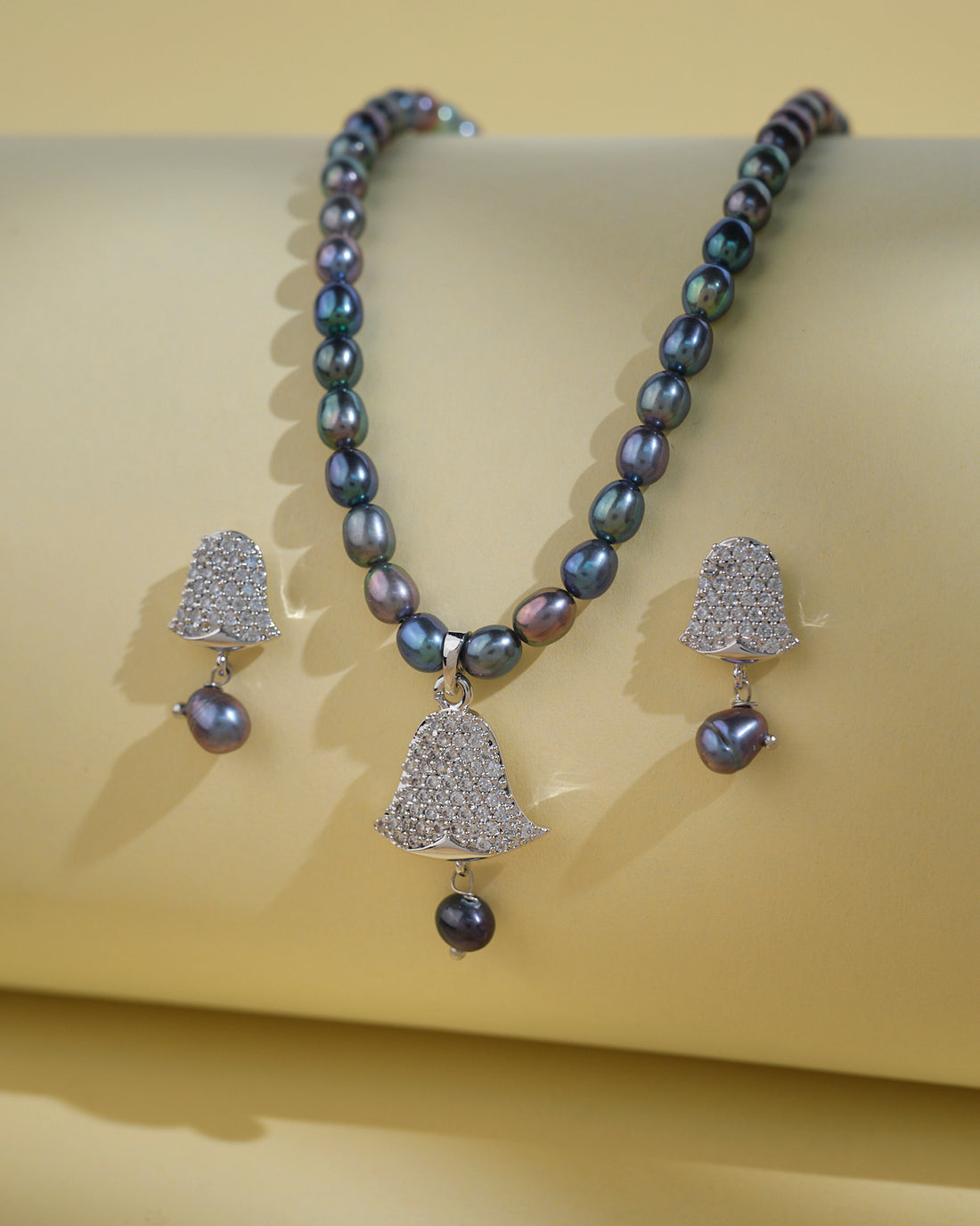 Chandrani Pearls India's Beautiful Pearl Necklace Sets feature a bell-shaped pendant and matching studded bell earrings with dangling pearls, offering timeless elegance. Displayed on a pale yellow surface.
