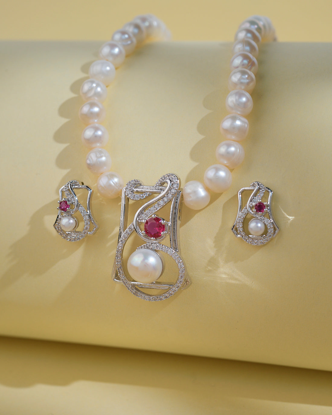The Trendy Pearl Necklace Set by Chandrani Pearls India, featuring a central diamond and pink gemstone pendant with matching pearl earrings, radiates timeless brilliance on a light yellow surface.