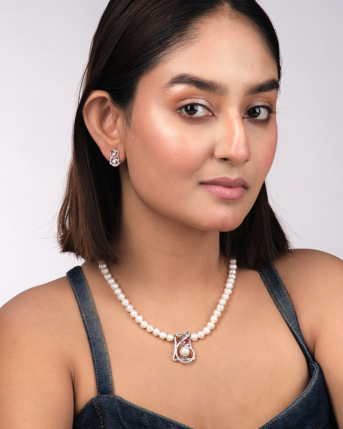 The Trendy Pearl Necklace Set by Chandrani Pearls India, featuring a central diamond and pink gemstone pendant with matching pearl earrings, radiates timeless brilliance on a light yellow surface.