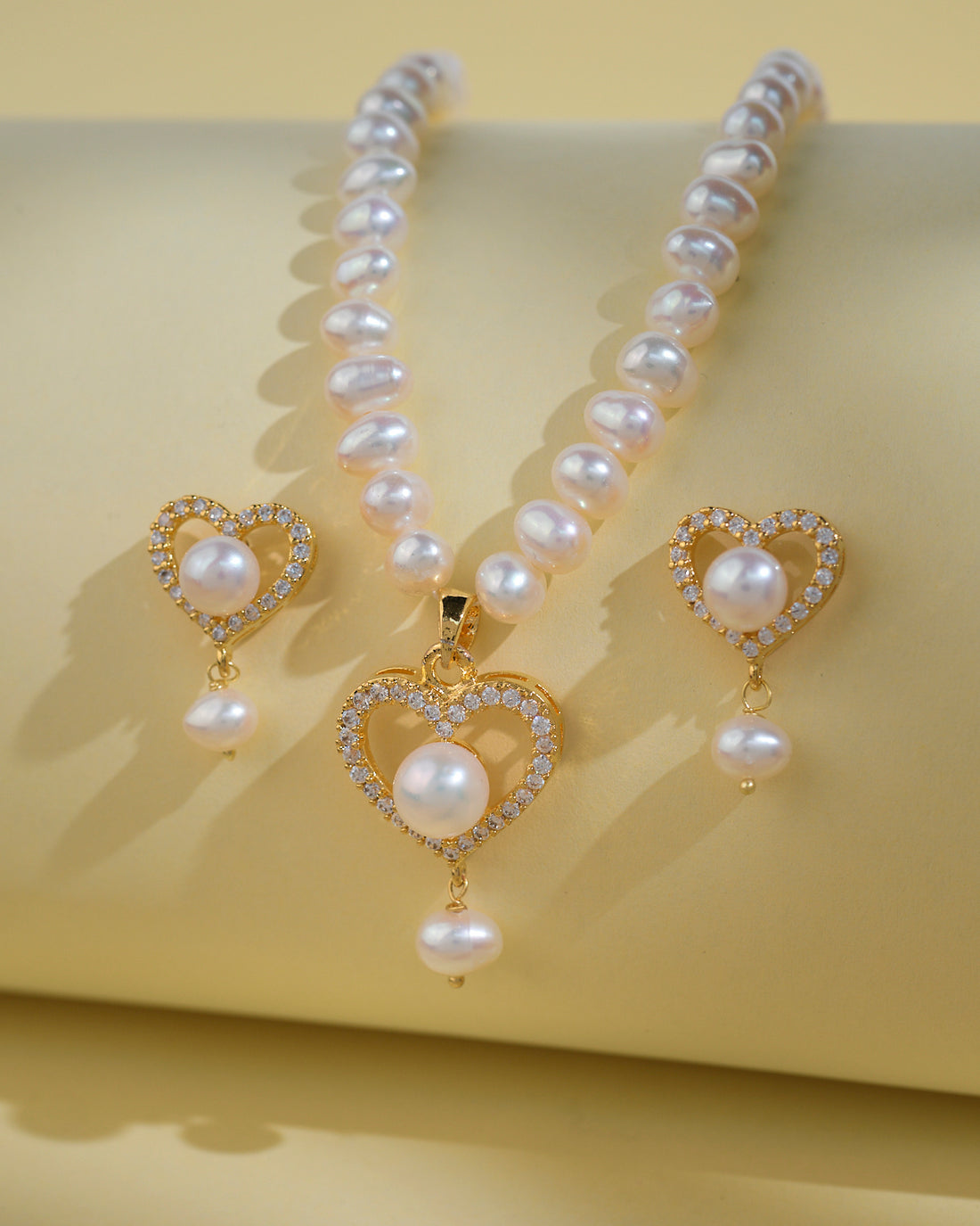 The Heart Pearl Necklace Set by Chandrani Pearls India features a pearl necklace with a gold heart-shaped pendant and matching earrings adorned with pearls, heart designs, and rhinestones, displayed on a light yellow surface.