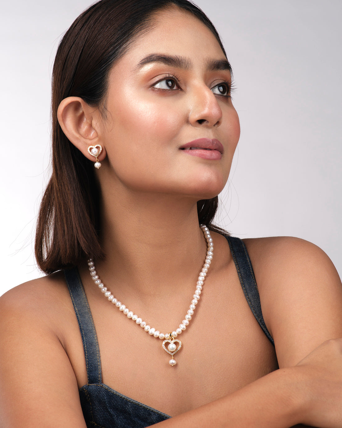 The Heart Pearl Necklace Set by Chandrani Pearls India features a pearl necklace with a gold heart-shaped pendant and matching earrings adorned with pearls, heart designs, and rhinestones, displayed on a light yellow surface.