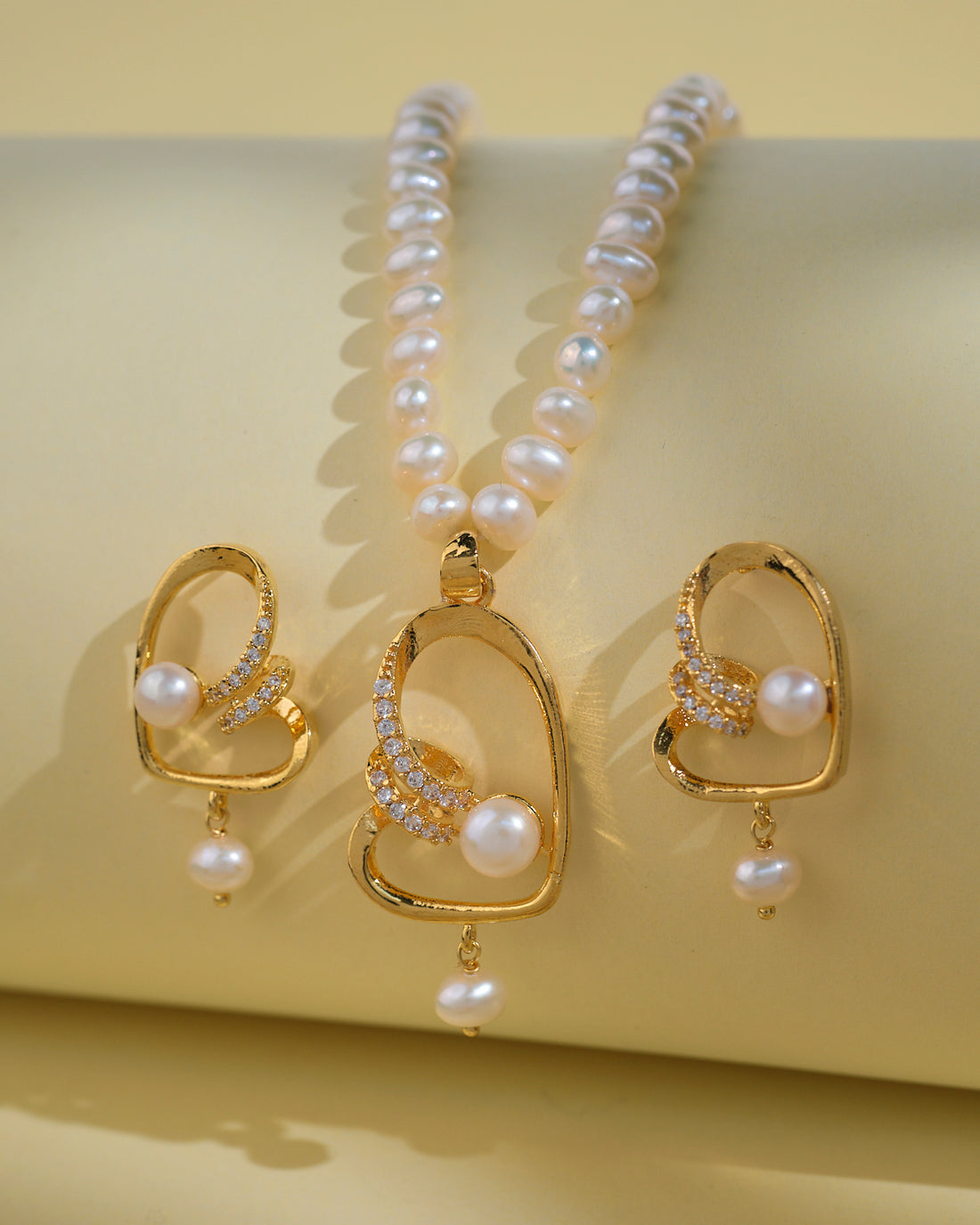 The Fashionable Pearl Necklace Sets by Chandrani Pearls India feature a gold-finish necklace and matching earrings with classic pearls and rhinestones, elegantly displayed on a light yellow background.