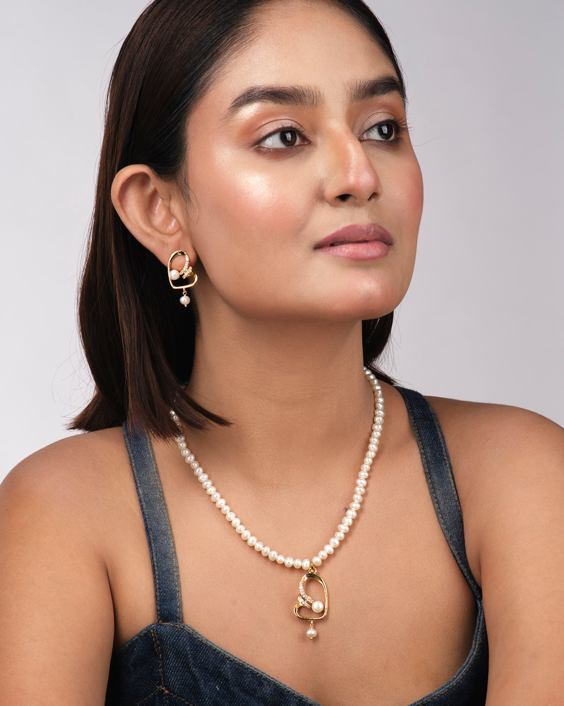 The Fashionable Pearl Necklace Sets by Chandrani Pearls India feature a gold-finish necklace and matching earrings with classic pearls and rhinestones, elegantly displayed on a light yellow background.