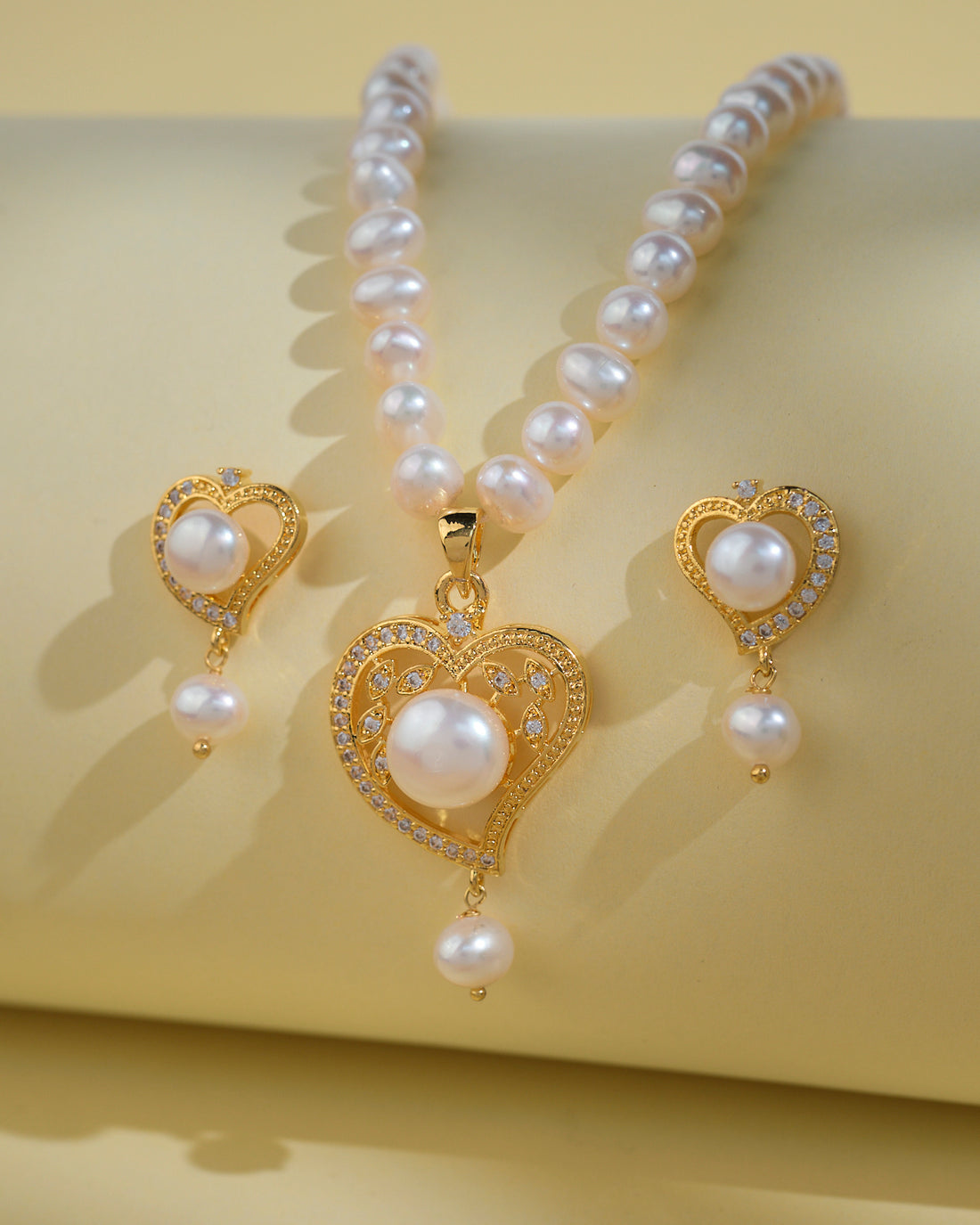Chandrani Pearls India Heart Pearl Necklace Sets: gold heart-shaped pearls with sparkling stones and matching earrings, elegantly displayed on a light yellow background—ideal for the modern woman.
