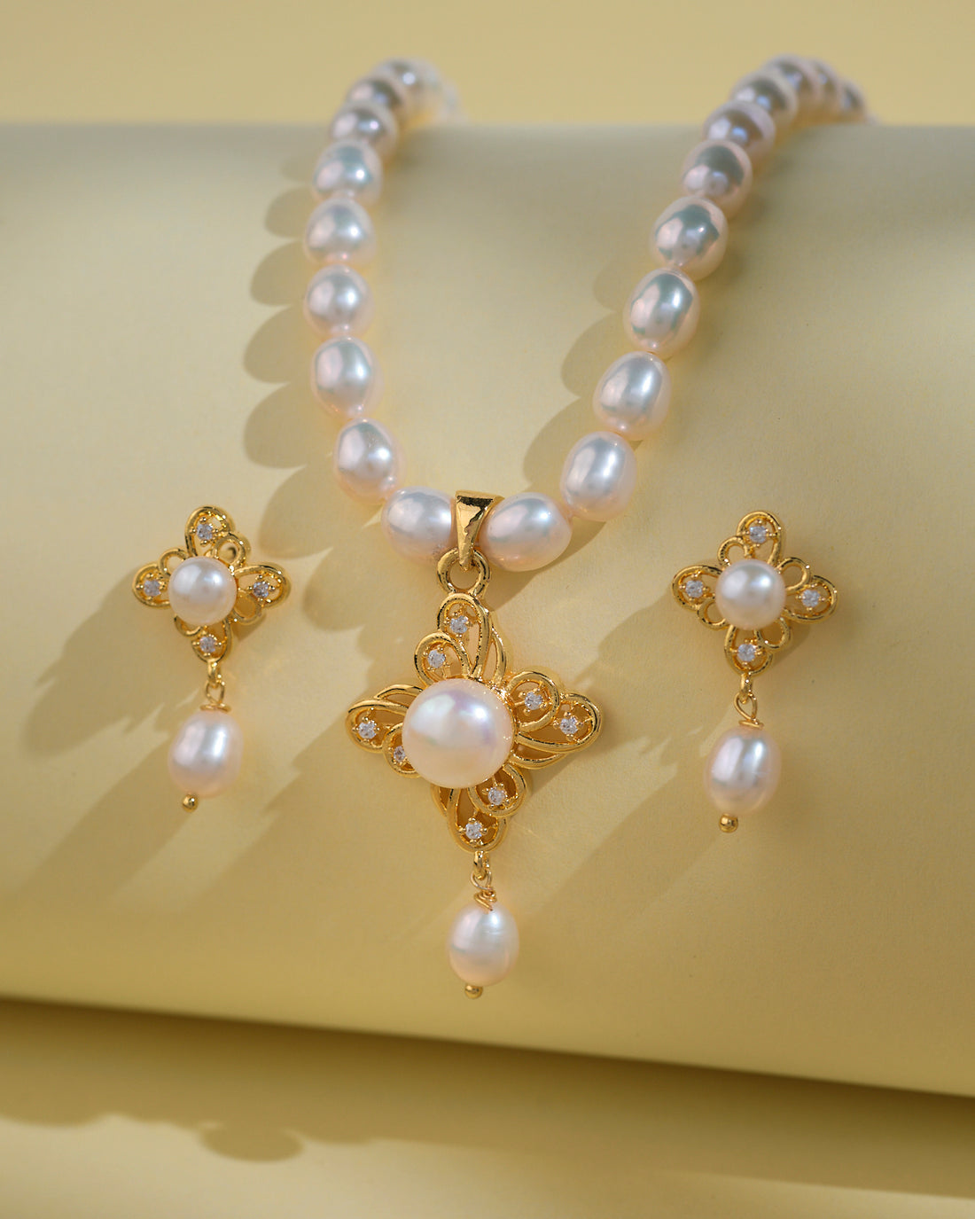 The Regal Pearl Necklace Sets by Chandrani Pearls India, featuring gold earrings with pearl and crystal accents, are showcased on a light yellow surface—an elegant choice for the modern woman.