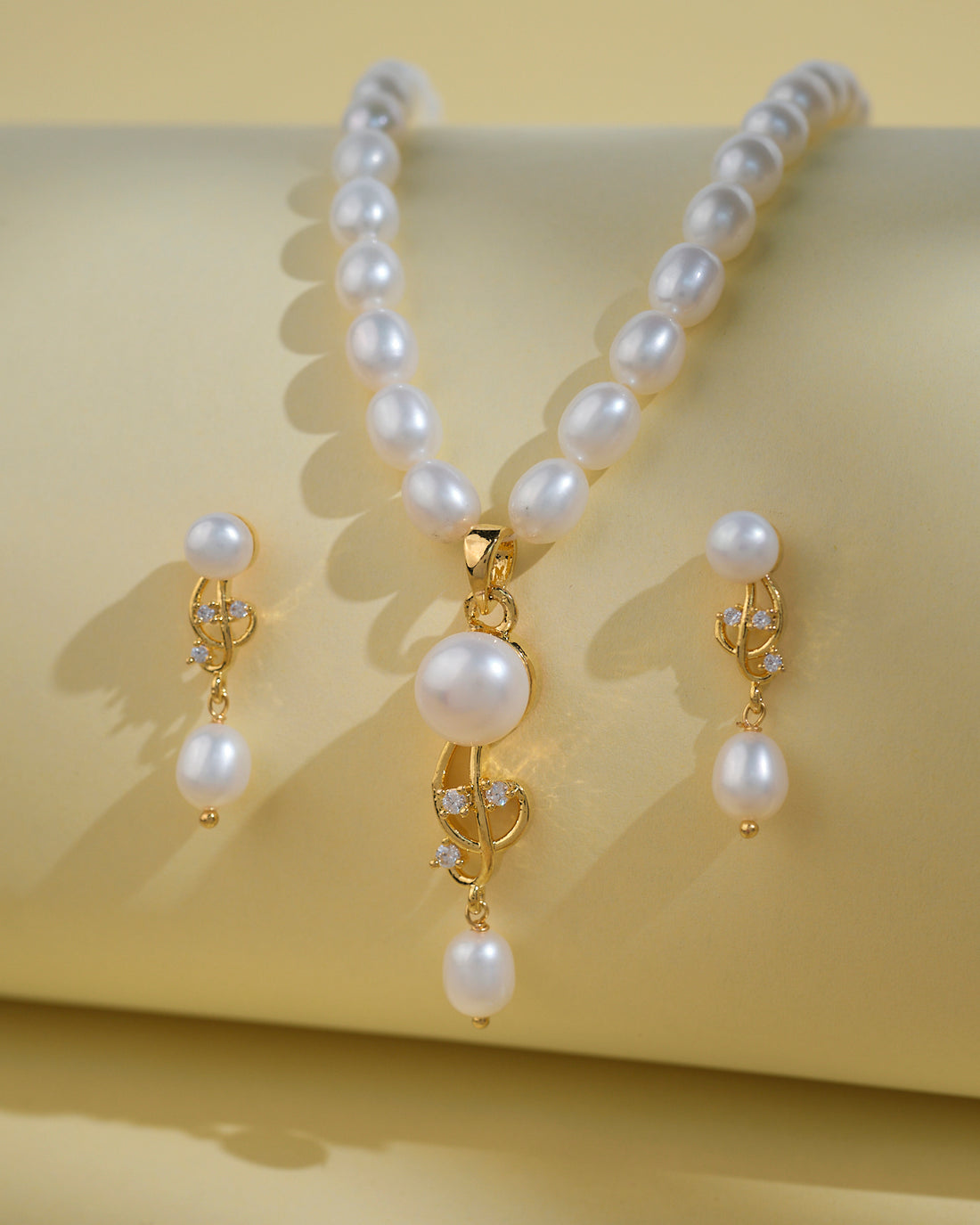 Chandrani Pearls India's Elegant Pearl Necklace Sets, featuring a gold and pearl pendant with matching drop earrings, are showcased on a pale yellow background—a sophisticated choice for today's modern woman.